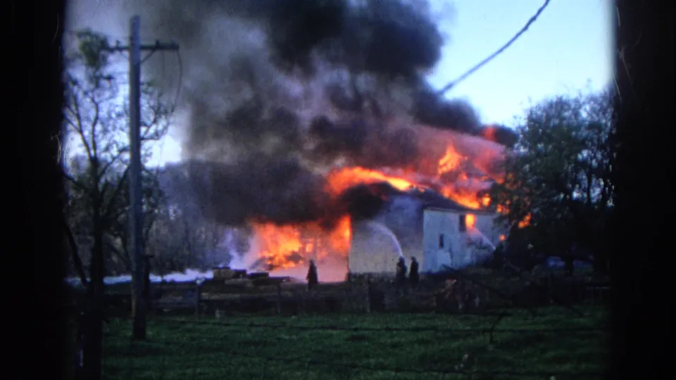1968 Wauconda, Illinois home-movie frame with firefighters silhouetted near a white building burning under thick smoke.