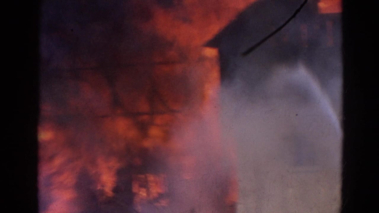 Flames and smoke fill a dark warehouse interior or wall opening as water spray crosses the frame.