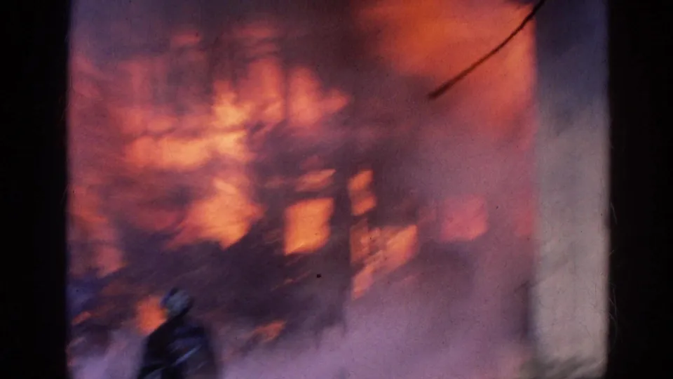 A firefighter in dark gear stands before the burning Wauconda warehouse as flames and smoke fill the 8mm frame.