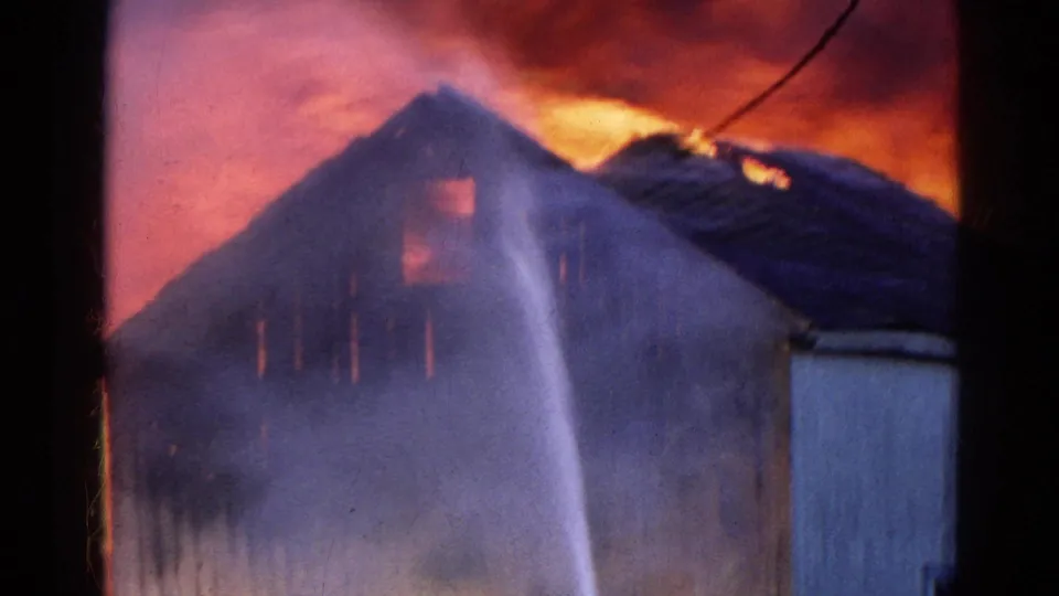 Firefighters direct water onto a burning warehouse in Wauconda, Illinois, with roof flames and smoke visible in 1968 8mm footage.