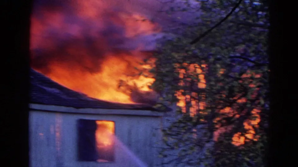 1968 Wauconda fire scene with flames over a house roof and a firefighter hose stream aimed into a burning window.