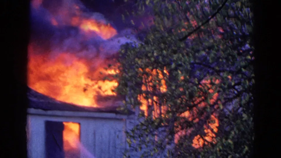 1968 Wauconda 8mm frame showing intense flames and smoke around a house, partially hidden by branches and a dark tree trunk.