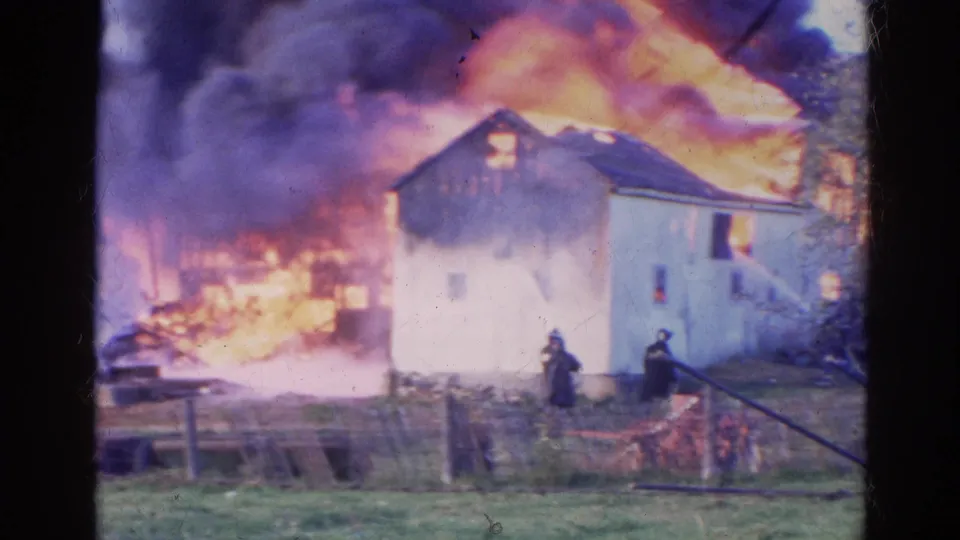 1968 Wauconda 8mm wide frame showing firefighters and hoses in a yard as a house and nearby structure burn.