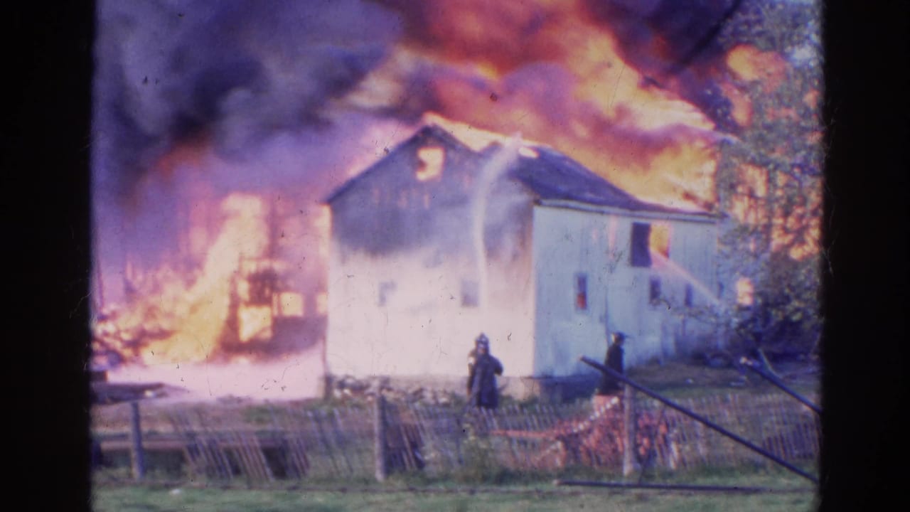 Firefighters spray water toward a light-colored house burning behind a wooden fence.