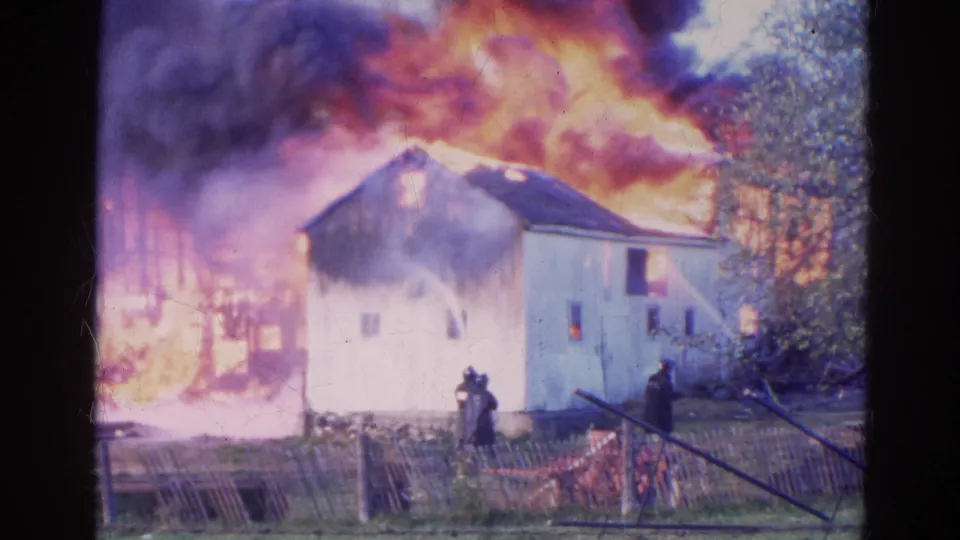 1968 Wauconda, Illinois home-movie frame with heavy flames behind a house and firefighters operating hoses from the yard.