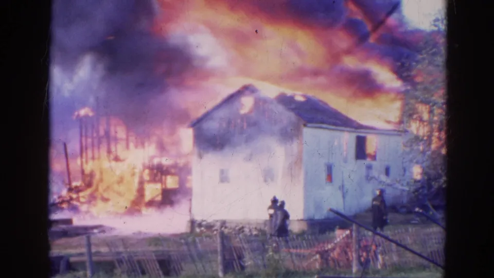 1968 Wauconda, Illinois frame showing a residential fire with flames behind the house, thick smoke, and firefighters in the foreground.