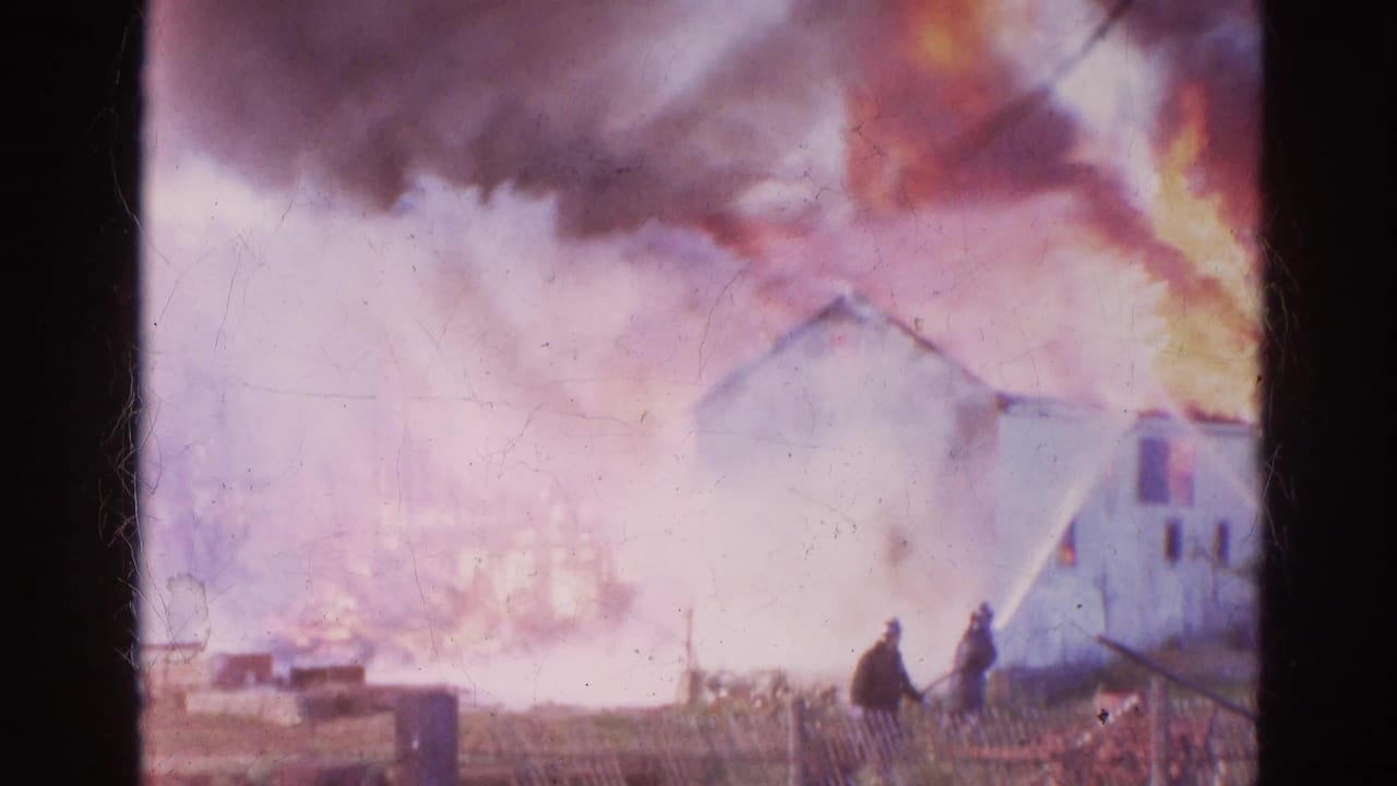 A distant view of the Wauconda fire shows two figures near a fence while smoke, flames, and water spray fill the frame.