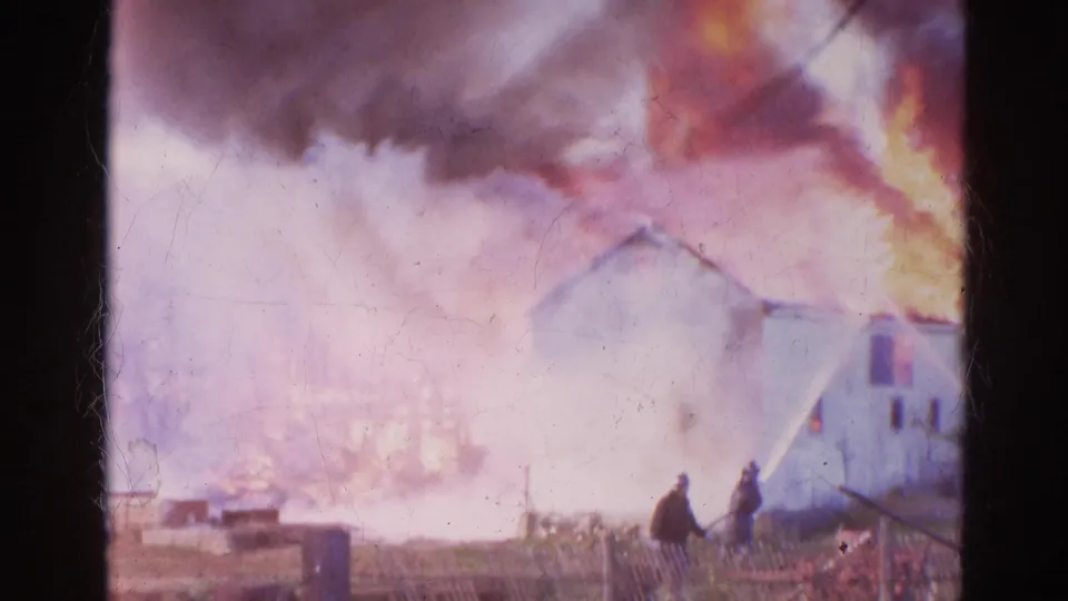 A distant view of the Wauconda fire shows two figures near a fence while smoke, flames, and water spray fill the frame.