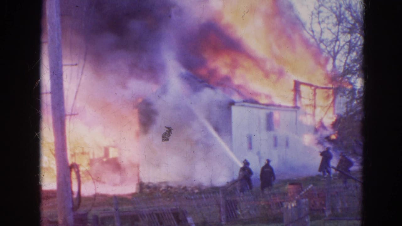 Firefighters spray water toward a white house burning behind smoke and flames.