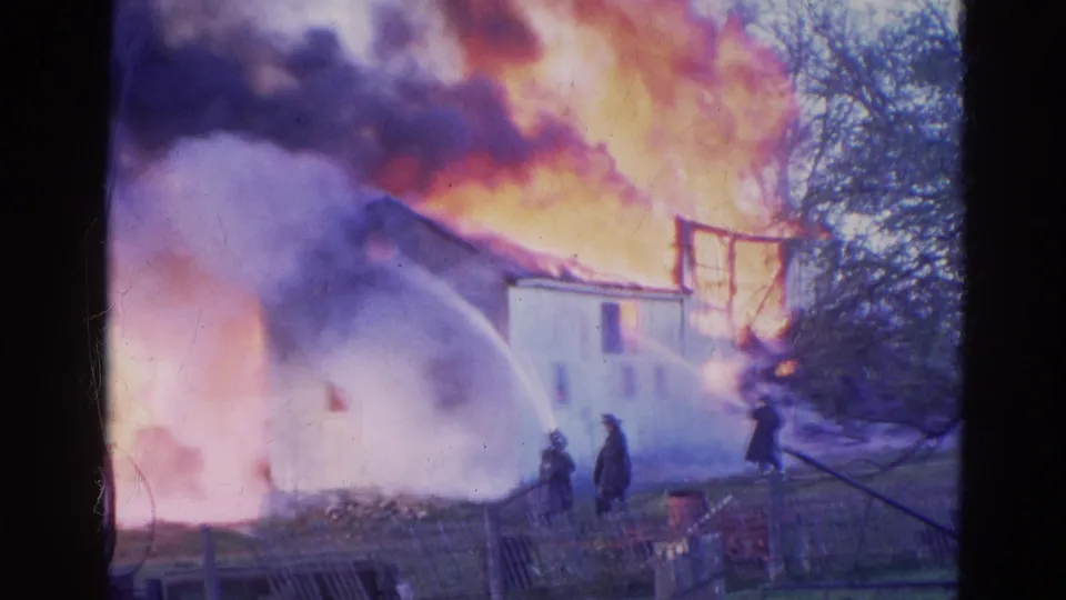 8mm footage frame from Wauconda, Illinois shows firefighters beside a white house obscured by smoke and active flames.
