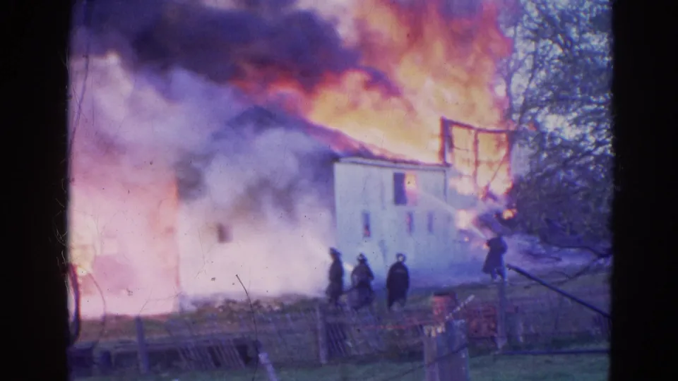1968 home-movie frame shows firefighters near a white house in Wauconda with flames and smoke rising above the structure.