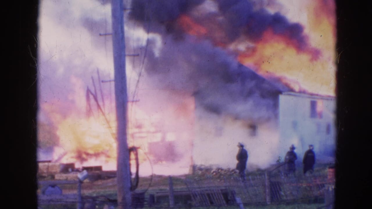 A utility pole stands before firefighters and bystanders near a house fire with heavy smoke.