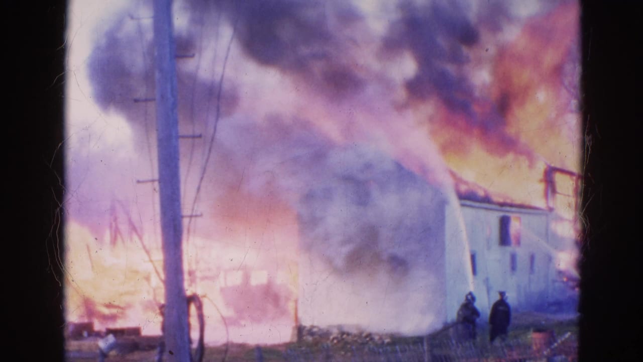 White building burning with flames, smoke, utility poles, and two people near the foreground.