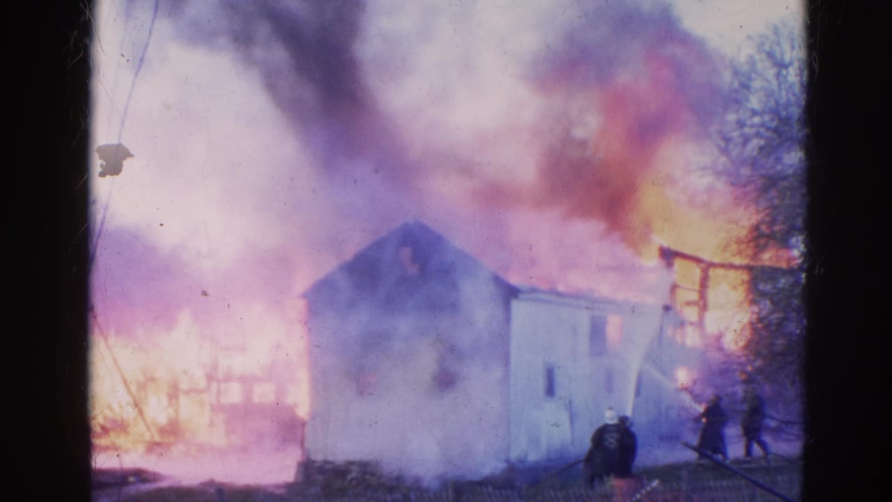 Firefighters stand near a small white house as flames and smoke rise behind it.