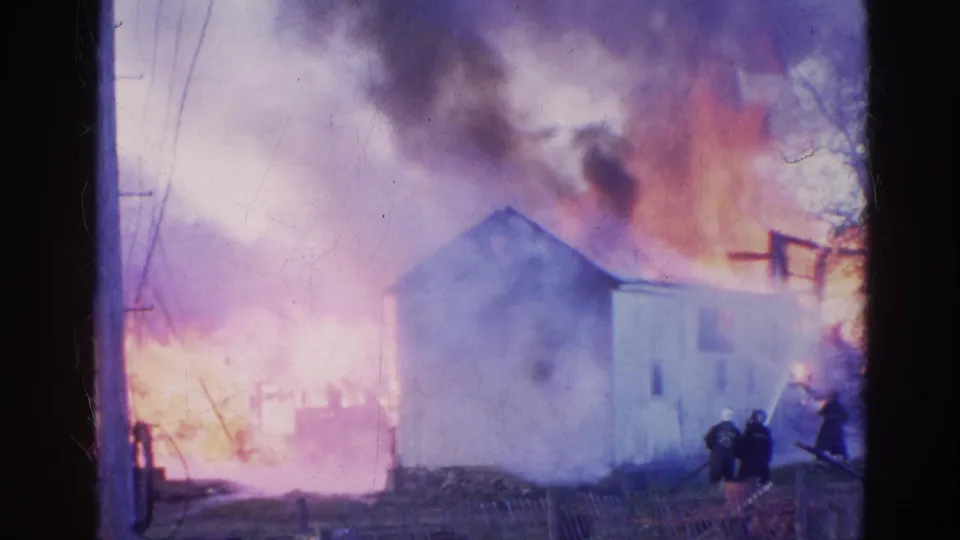 A small white structure in Wauconda, Illinois is partly obscured by smoke as firefighters stand near active flames in this 1968 8mm footage.
