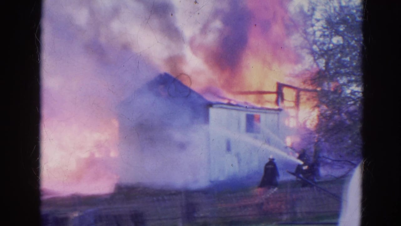 White building partly obscured by smoke and flames, with a firefighter or onlooker near a hose in the foreground.