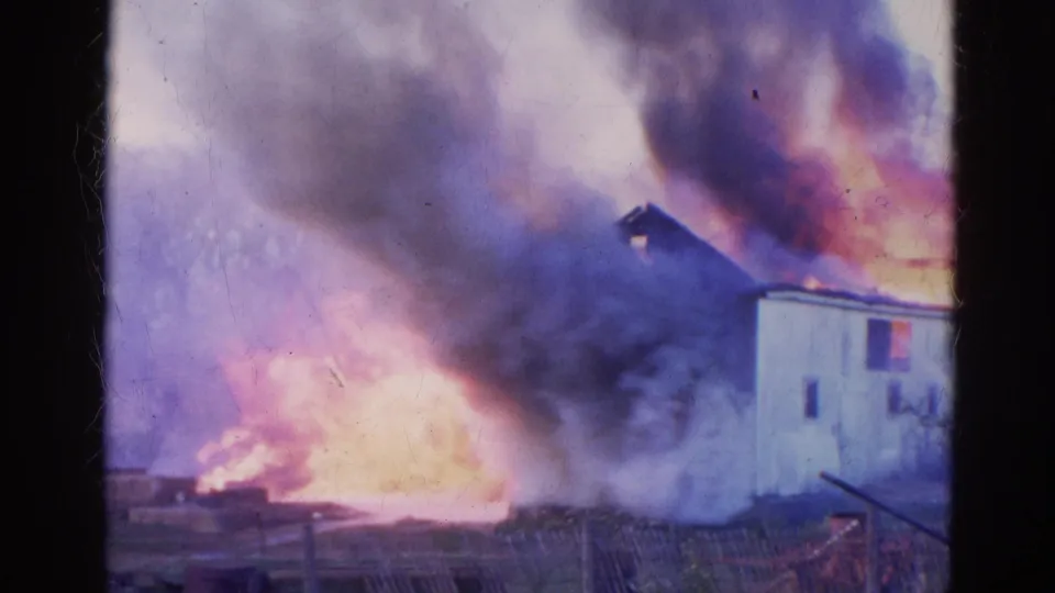 1968 Wauconda frame with bright flames and thick smoke spreading beside a white building, seen across a low fence.