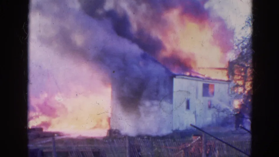 1968 Wauconda 8mm frame showing thick smoke and orange fire surrounding part of a white building behind a fence.
