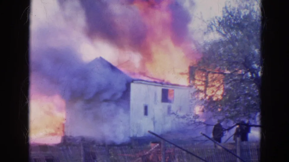 1968 Wauconda frame showing a white building amid flames and smoke, with two distant onlookers or responders near the fence.