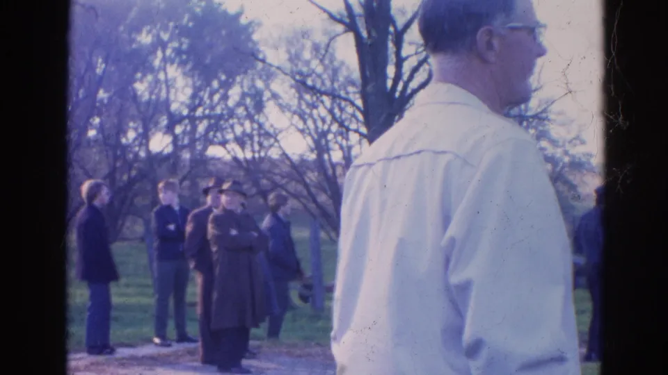 Bystanders in Wauconda, Illinois stand near trees and a wooden fence as a man in a light shirt and glasses appears in the foreground.