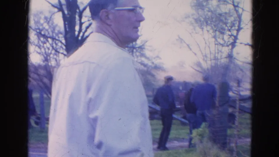 A foreground man in glasses stands in profile while other adults near a fence watch the smoky fire scene in 1968 Wauconda, Illinois.