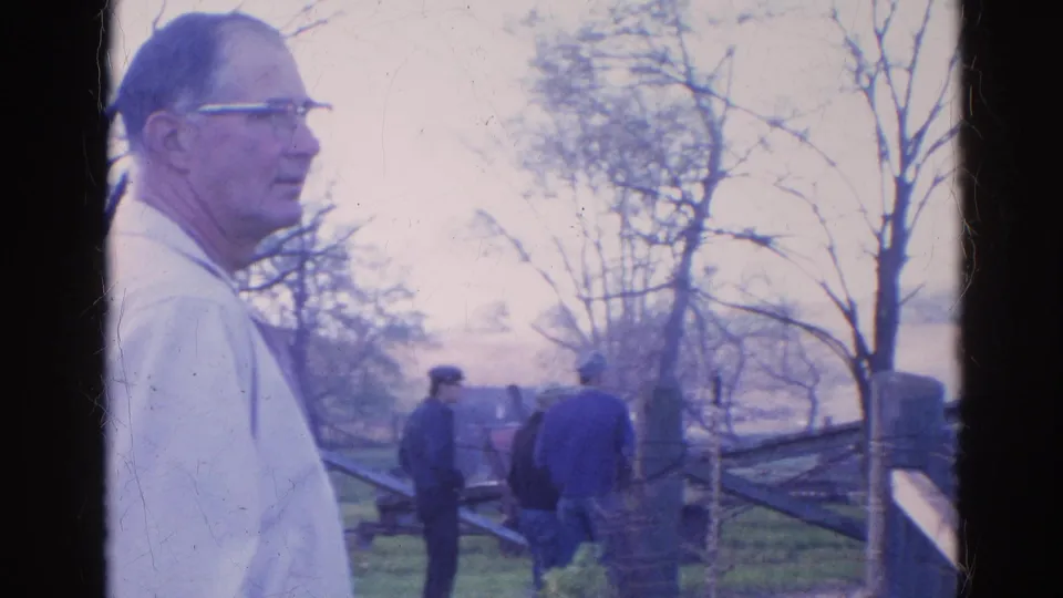 A man in a light shirt and glasses stands close to camera as two men by a wooden fence look toward smoke from the Wauconda fire.