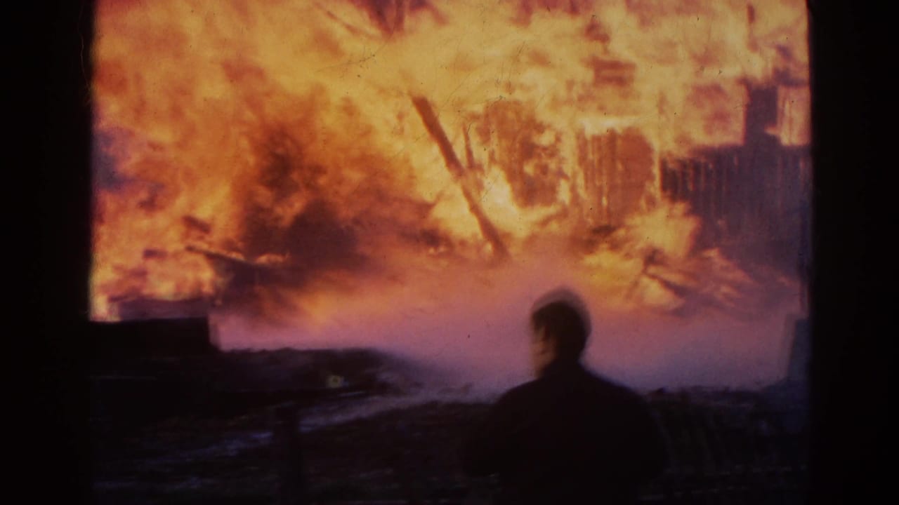 A silhouetted person watches a building engulfed by tall orange flames.