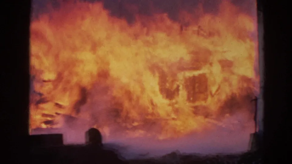The 8mm footage shows the building fire at peak brightness, with flames washing across the structure and a viewer partly visible below.