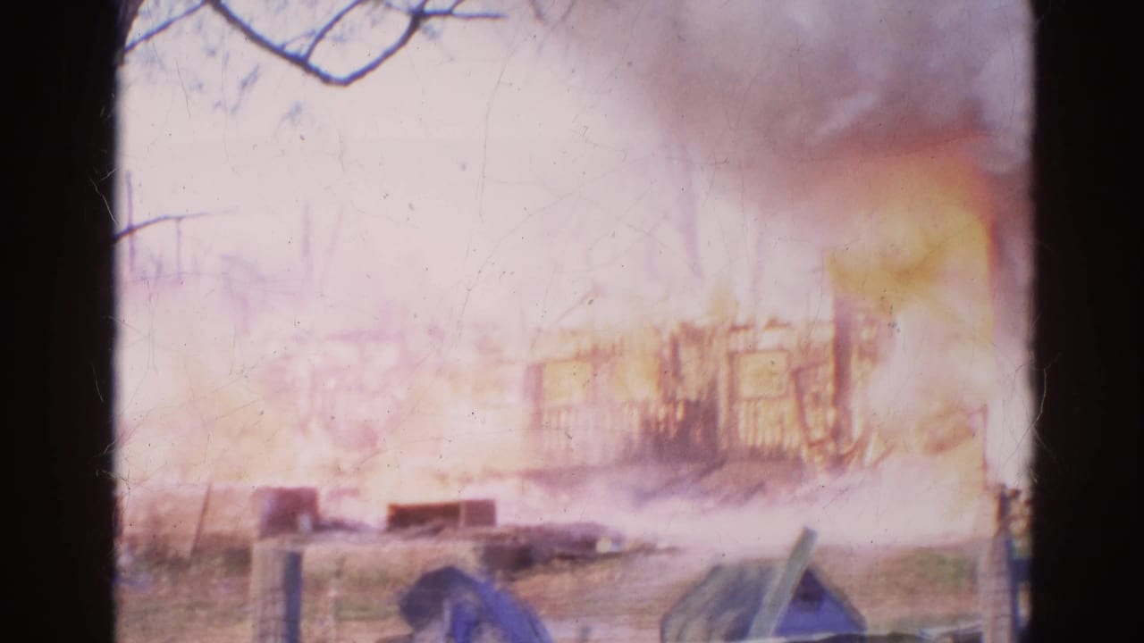 1968 Wauconda 8mm frame showing a wooden structure engulfed by flames with smoke rising behind bare branches and roadside foreground elements.
