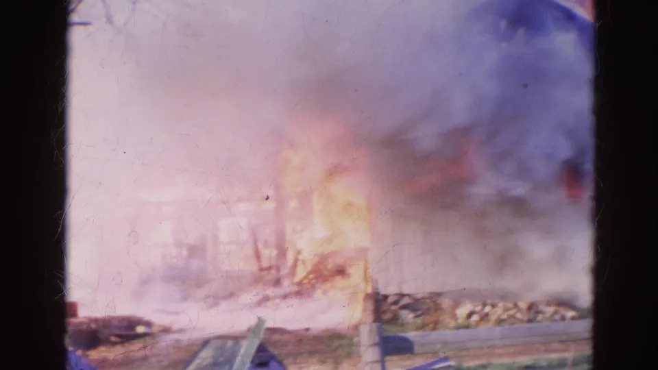 1968 Wauconda 8mm frame showing smoke, flames, ground debris, and a low roadside view of a burning wooden structure.