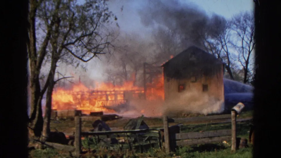 The 1968 8mm frame shows a rural structure fire near a small building, with smoke drifting above fencing and trees.