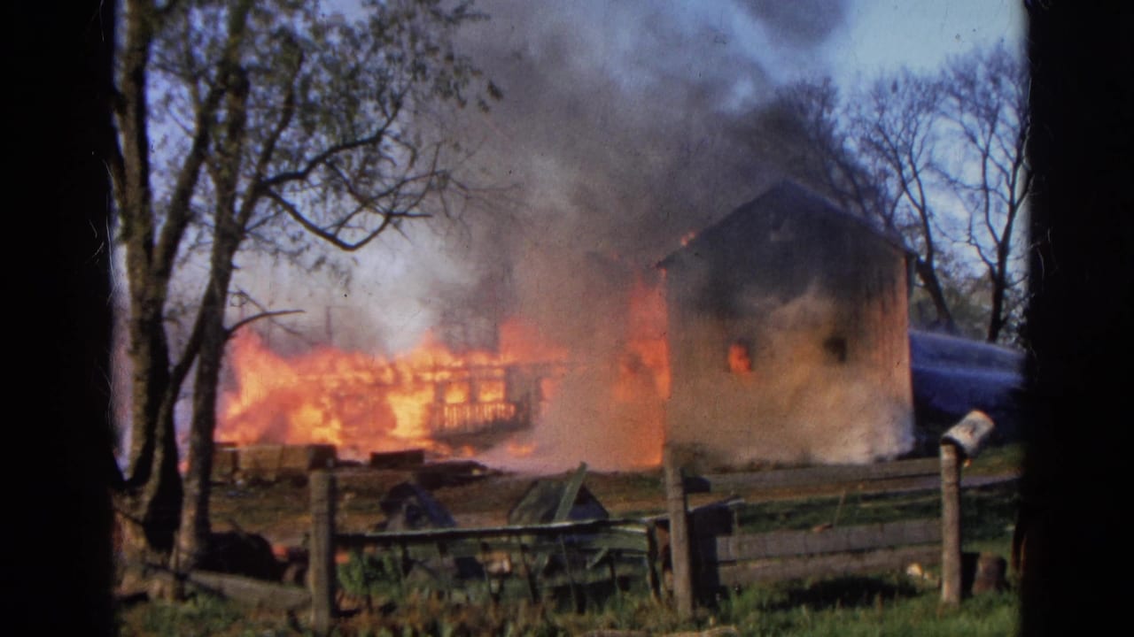 Burning rural building with flames, smoke, trees, and wooden fence in the foreground.