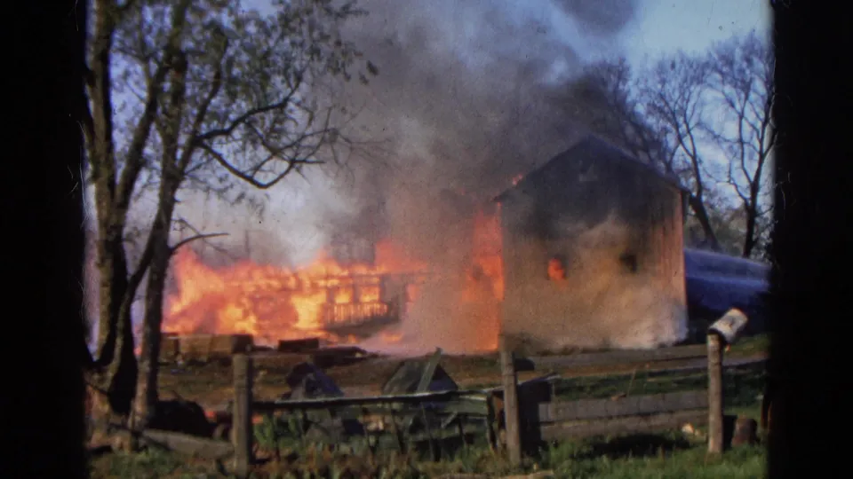 1968 Wauconda, Illinois 8mm frame showing a rural building engulfed in flames behind a wooden fence and bare trees.
