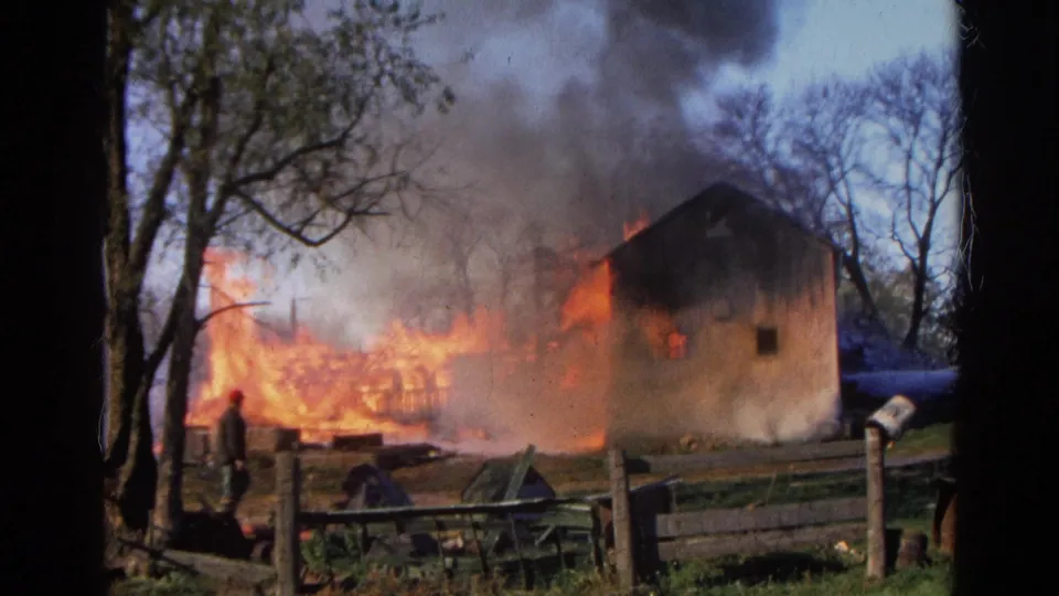1968 Wauconda, Illinois frame with an adult observer near a fence as fire consumes a rural structure.