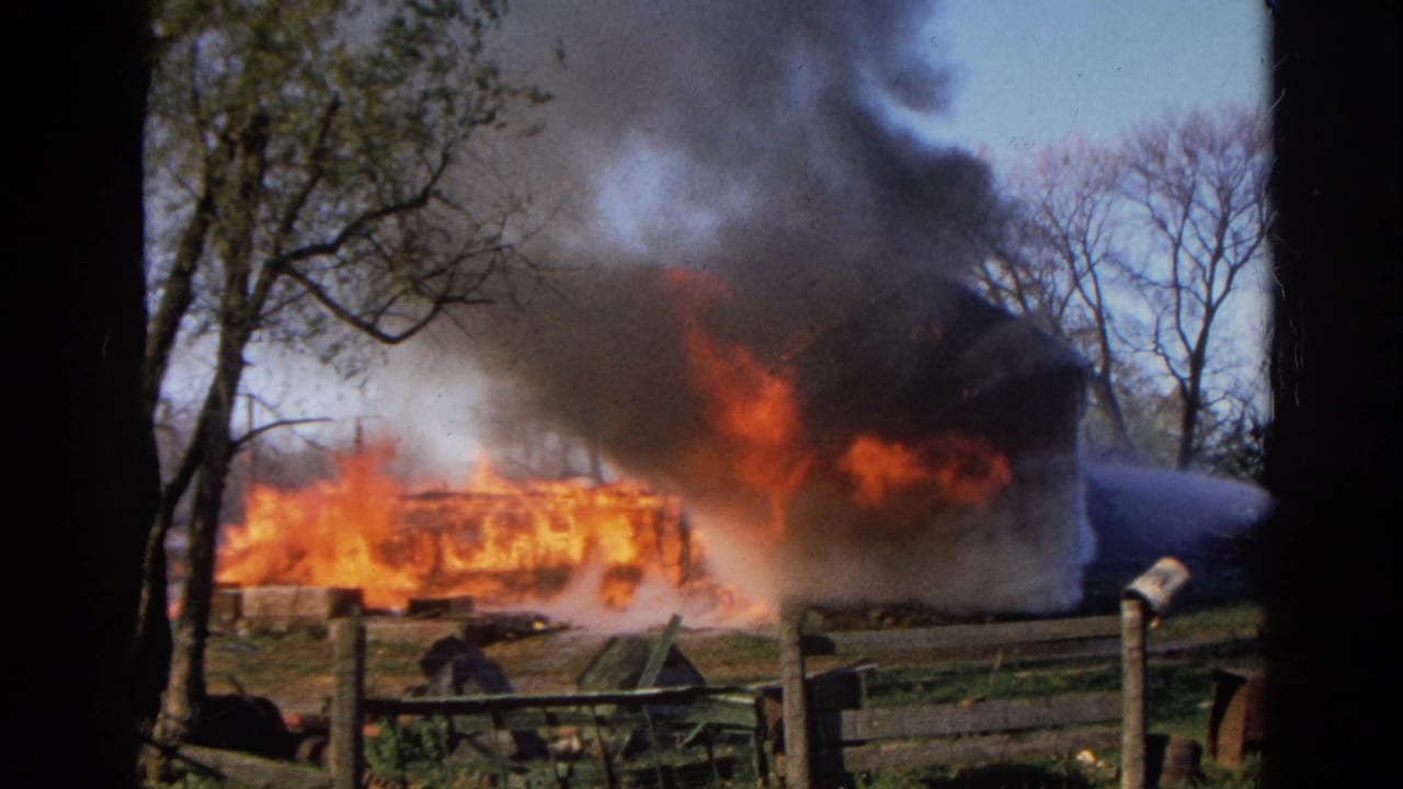 Barn or outbuilding burning behind a fence as smoke rises over trees.