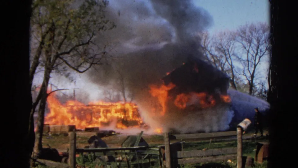 1968 Wauconda, Illinois frame with a firefighter on the right spraying water at a burning barn or outbuilding amid heavy smoke.