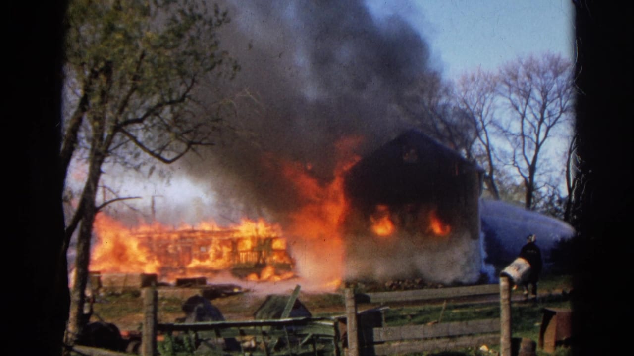 1968 Wauconda, Illinois 8mm frame showing a structure engulfed in flames with dark smoke, trees, fencing, and firefighting water spray.
