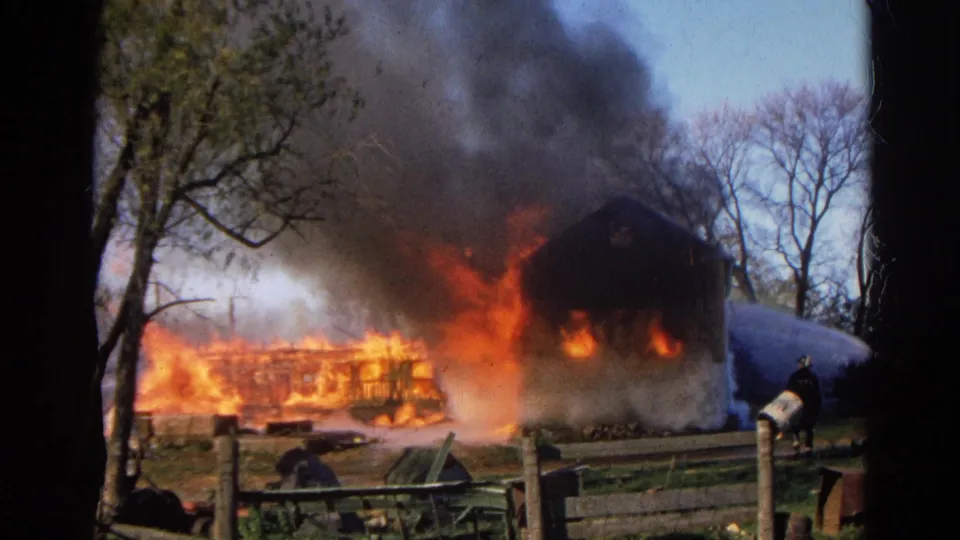 1968 Wauconda, Illinois 8mm frame showing a structure engulfed in flames with dark smoke, trees, fencing, and firefighting water spray.