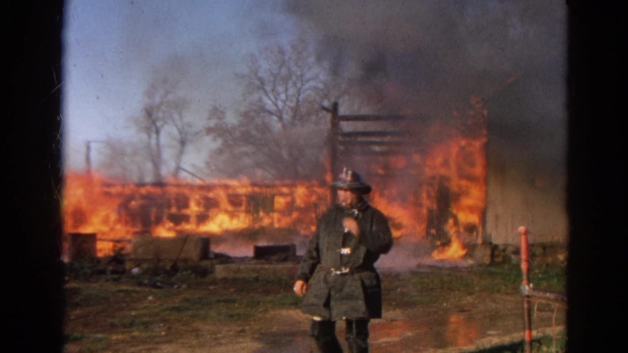 Firefighter stands near a large fire with smoke, flames, hose spray, and charred framing behind him.
