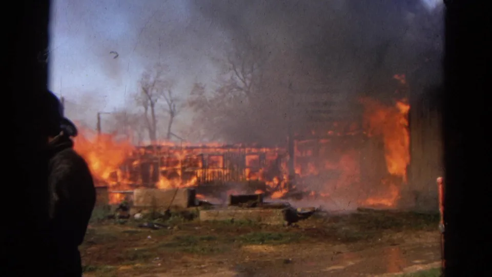 A rural barn burns fiercely in the background while a dark figure stands partly obscured near the center of the 8mm frame.