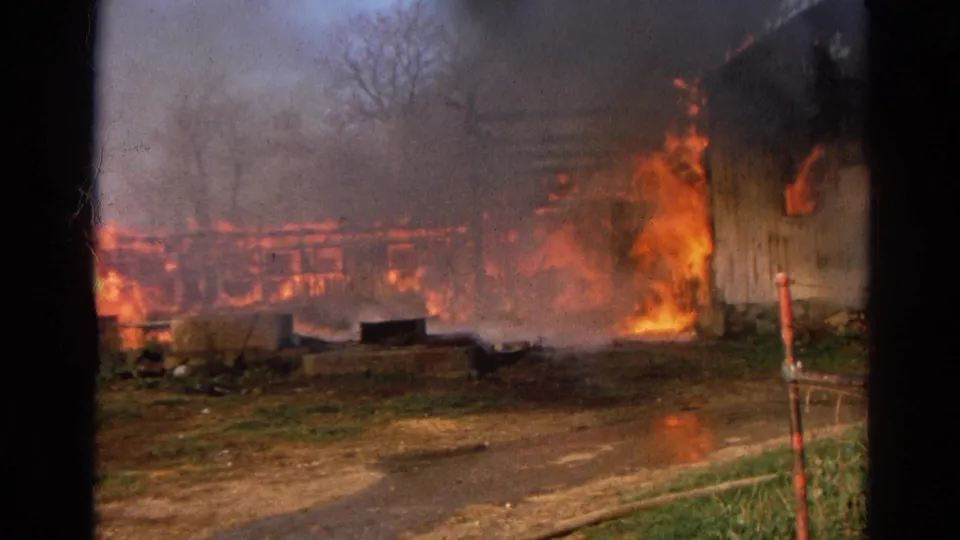 The 1968 Wauconda barn fire fills the frame, with flames climbing the wooden wall and smoke softening the rural background.