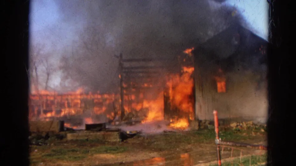 A rural wooden barn in Wauconda, Illinois burns intensely, with charred framing, heavy smoke, and firelight visible on wet ground.