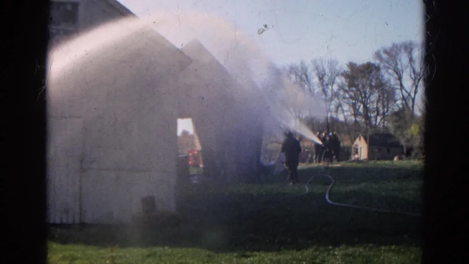 1968 Wauconda, Illinois frame showing a firefighter and hose line near a smoking farm building with water spray crossing the scene.