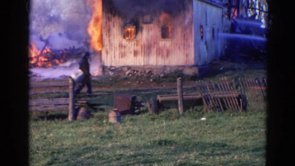 1968 Wauconda, Illinois frame showing a man in a dark coat and hat walking near a burning barn, with smoke and a fence in the foreground.