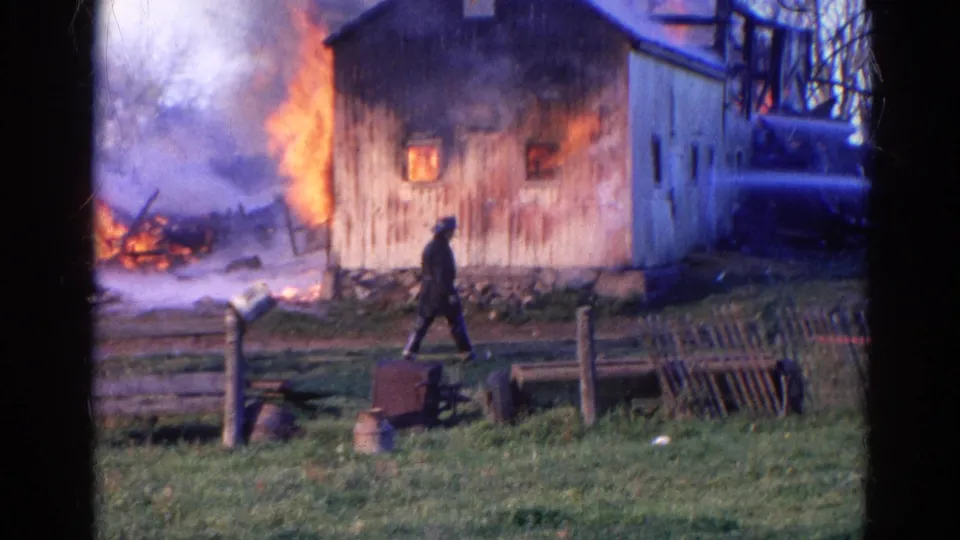 1968 Wauconda, Illinois 8mm frame with a man walking in front of a barn fire, smoke behind the building, and a possible fire vehicle at right.