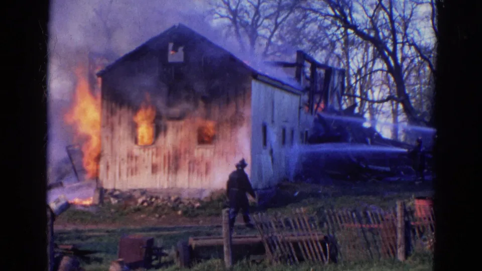 1968 Wauconda, Illinois frame showing the barn still burning, heavy smoke above it, and a dark-clad man near the foreground fence.