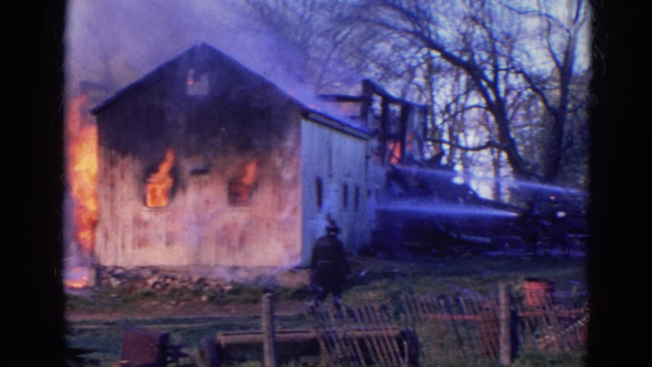 Barn burning beside trees with flames at the wall and a firefighter near a fence.