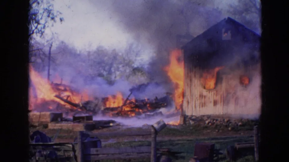 A handheld 8mm view shows heavy smoke, active flames, burned debris, fencing, and the side of the barn during the rural fire.