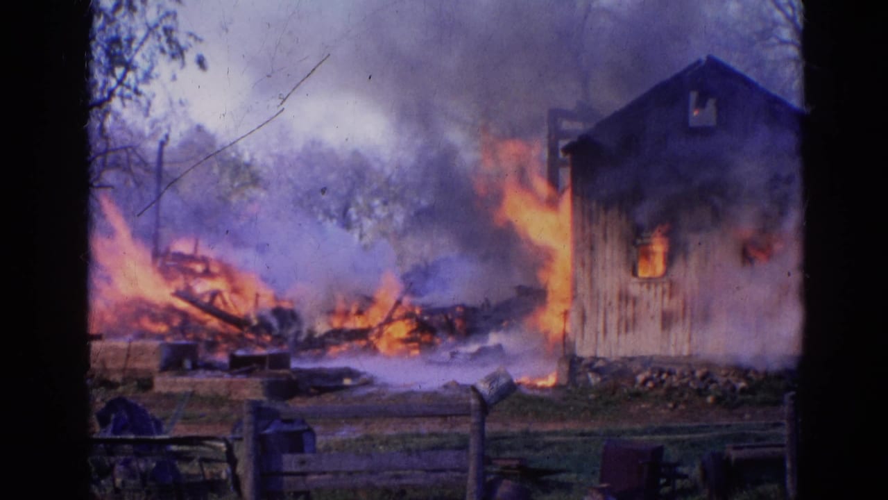 1968 Wauconda, Illinois 8mm frame showing flames consuming debris beside a wooden structure as smoke rises behind a foreground fence.