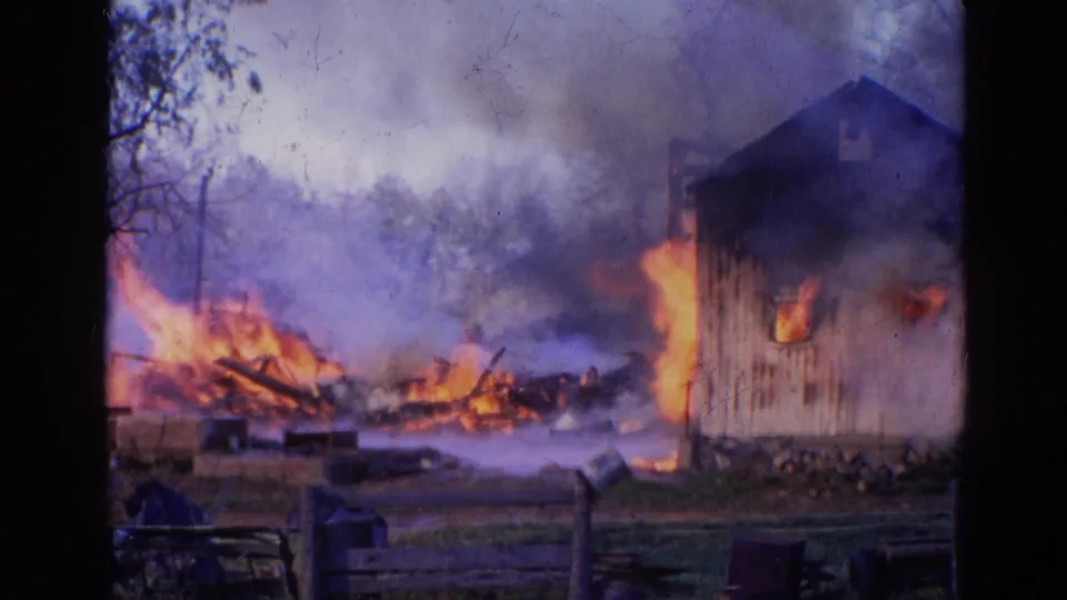1968 Wauconda, Illinois home-movie frame with a wooden building burning at right and flames moving through debris behind a fence.